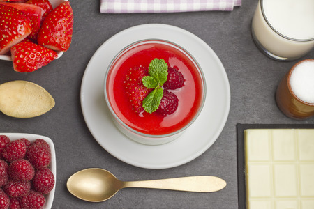 White chocolate panna cotta with raspberries and strawberries on a slate plate with ingredients, a golden spoon and a purple napkinの写真素材