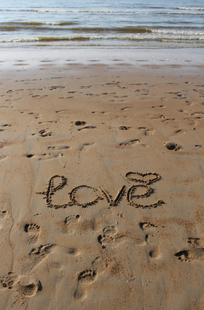 Inscription of "Love" and a symbol of heart in the sand. Footprints in the wet sand and sea waves.の写真素材