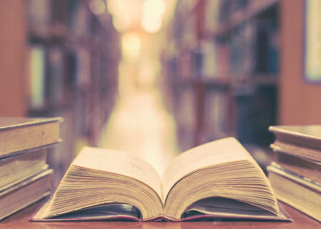 Old book in library with open textbook, stack piles of literature text archive on reading desk, and blur aisle of bookshelves in school study class room background for education learning conceptの写真素材