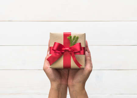 Christmas gift box with red satin ribbon and spruce twig decoration on giver hands giving out on white pine wood table background for boxing day, winter seasonal holiday and new year celebrationの写真素材