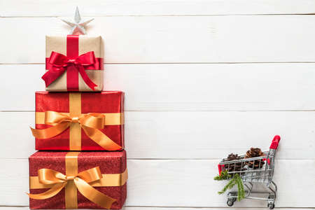 Gift box present stack in Christmas tree shape with red-gold ribbon, star, spruce twig, pine cone and shopping trolley on white wood for Xmas boxing day winter holiday seasonal celebration backgroundの写真素材