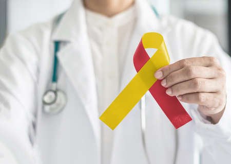 World hepatitis day and HIV/ HCV co-infection awareness with red yellow ribbon in medical doctor hand symbolic bow color to support patient with illness and hepatic diseaseの写真素材