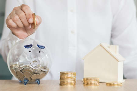 Home loan, banking, money saving and financial planning concept with piggy bank jar, house bank with person's hand inserting gold coin into glass piggybankの写真素材