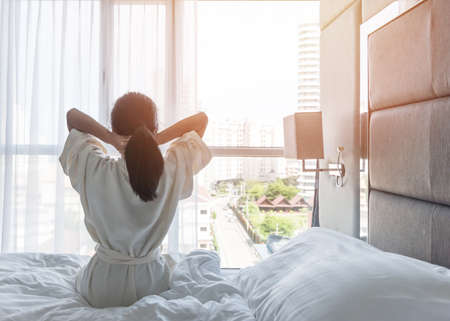 Easy lifestyle Asian woman waking up from good sleep in weekend morning taking some rest, relaxing in comfort bedroom at hotel window, having happy lazy day enjoying work-life quality balance conceptの写真素材
