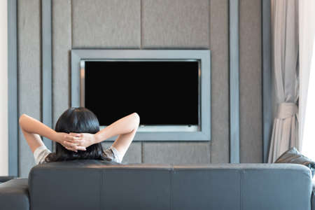 Relaxing at home concept with girl sitting on couch watching television in luxurious house or hotel interior living room (with blur blank TV screen background)の写真素材