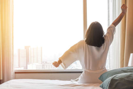 Easy lifestyle Asian woman waking up from good sleep in weekend morning taking some rest, relaxing in comfort bedroom at hotel window, having happy lazy day enjoying work-life quality balance conceptの写真素材