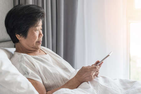 Elderly senior woman aged person lifestyle using mobile tablet and digital internet technology reading e-book, social media network on bed in nursing home or health careの写真素材