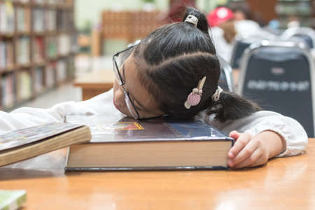 Asian kid student studying hard feeling tired in school library sleeping on big book in library or...