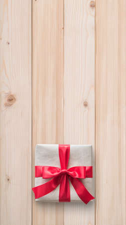 Gift box present (isolated with clipping path) with red bow satin ribbon over brown wrapping paper on white pine wood background for Christmas and birthday greeting card design decorationの写真素材