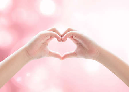Heart-shape hand gesture of kid's body language for children's love, peace, kindness and world humanitarian aid concept. (Hand isolated on sky background with pink bokeh and sun flare)の写真素材