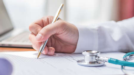 Doctor writing on medical health care record, patients discharge, or prescription form paperwork in hospital clinic office with physician's stethoscope on deskの写真素材