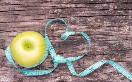 Slim diet, healthy eating habit for heart health and anti-aging concept with measuring tape wrapping around green apple on old aged wood backgroundの写真素材