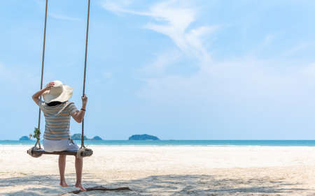 Summer sandy beach holiday vacation with young girl relaxing sitting on swing enjoying tropical ocean view, beautiful blue sunny sky on sea shoreの写真素材