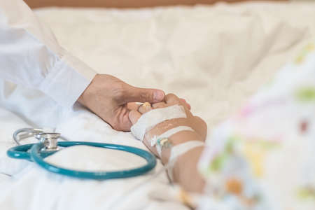 Nursing caretaker concept with kid patient sleeping in bed with pediatric doctorâs hand support in medical hospital background (focus on hand)の写真素材