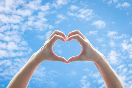 Woman hands in heart shape raising against blue sky with clouds for world kindness day, charity donation, friendship and Valentines love conceptの写真素材