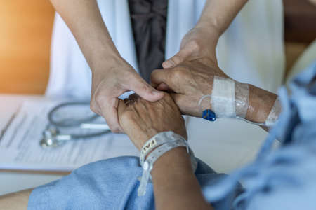 Elderly senior aged patient on bed with geriatric doctor holding hands for trust and nursing health care, medical treatment, caregiver and in-patient ward healthcare in hospitalの写真素材