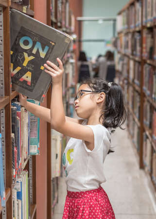 Student girl kid in school library selecting books for reading in class room from bookshelf 