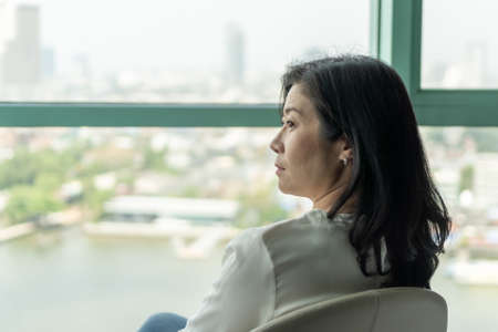 Middle-aged woman with modern living lifestyle, happy life quality concept with mature menopause female relaxing sitting in rest  hotel guest room or luxury home room with beautiful city viewの写真素材