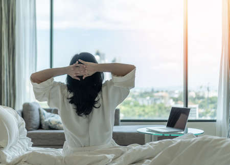 Work-Life balance and life quality concept with woman rear view in rest relaxing on bed, take it easy in modern hotel guest room or luxury home living room with beautiful business city urban sceneの写真素材