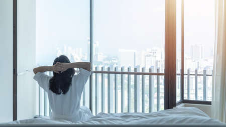 Easy lifestyle Asian woman waking up from good sleep in weekend morning taking some rest, relaxing in comfort bedroom at hotel window, having happy lazy day enjoying work-life quality balance conceptの写真素材