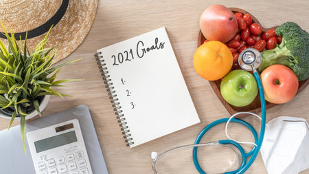 2021 goals on note book for new year list reminder in notebook of yearly planner and health plan for work-life balance on desk background of computer, healthy food, face mask for covid-19 protectionの写真素材