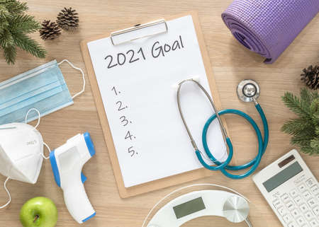 2021 new year goals in new normal lifestlye in health, covid-19 safety, weight loss, food diet, calories counting control and exercise written in white paper on medical note pad clipboard on work deskの写真素材