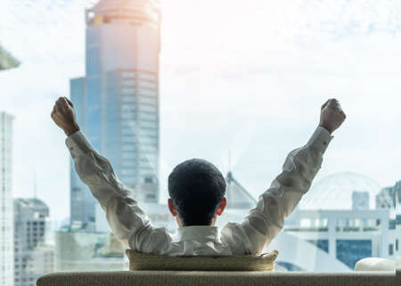 Business achievement concept with happy businessman relaxing in office or hotel room, resting and raising fists with ambition looking forward to city building urban scene through glass windowの写真素材
