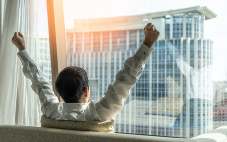 Business achievement ambition concept with businessman raising fists happily, relaxing in office or hotel room, resting and looking forward to city building urban sceneの写真素材