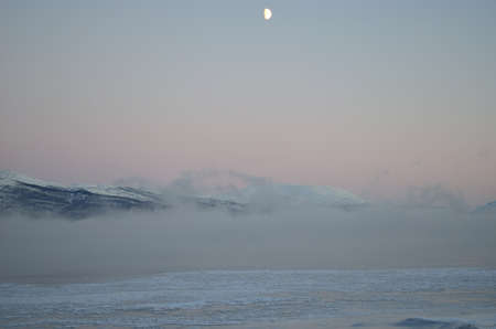 beautiful icy fjord covered in thick ice fog with moon and orange vibrant dawn sky and snowy mountain in the distanceの写真素材