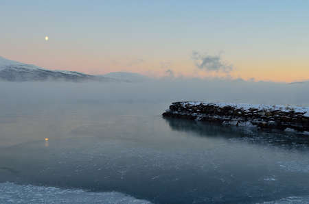 beautiful icy fjord covered in thick ice fog with moon and orange vibrant dawn sky and snowy mountain in the distanceの写真素材