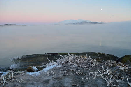 Stone pier in front with ice fog hovering over the sea and salt water frozen to ice by the shore...