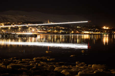 icy sea shore and beautiful fjord with city lights reflecting on the sea surface and airplane approaching for landing at nightの写真素材