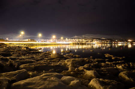 icy sea shore and beautiful fjord with city lights reflecting on the sea surface at nightの写真素材