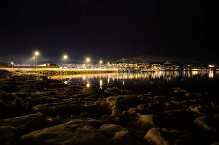 icy sea shore and beautiful fjord with city lights reflecting on the sea surface at nightの写真素材