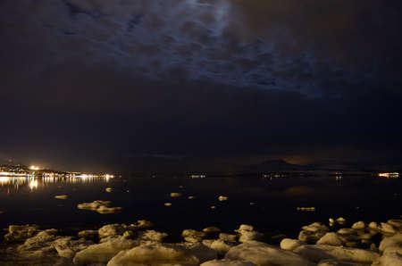 icy sea shore and beautiful fjord with city lights reflecting on the sea surface at nightの写真素材
