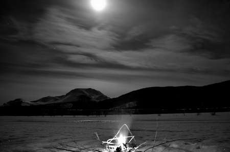 burning fire on frozen river bed with majestic snowy mountain and full moon in the backgroundの写真素材