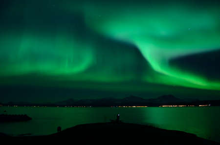 strong aurora borealis over fjord and mountain with tourist in frontの写真素材