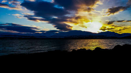 Golden sunny sunset over snowy mountain range with reflection on sea surface