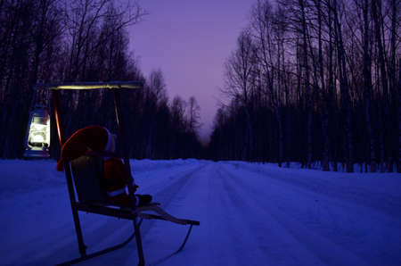 small santa travelling home on kicksled on snowy road with suitcase and lanternの写真素材
