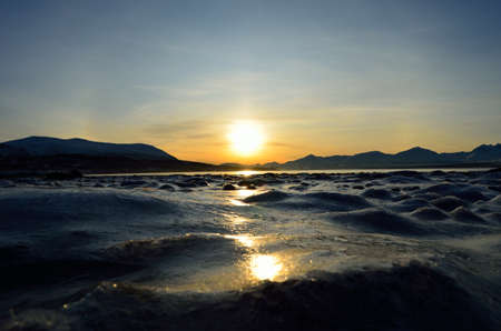 golden sunrise over blue fjord and snowy mountain with reflection on thick frozen sea shore ice