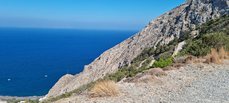 A person stands on a rocky hillside overlooking the sea. Waves gently lap at the shore. Small boats can be seen in the distance. The sky is clear and blue.の写真素材