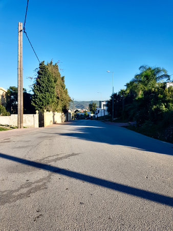 Asphalt road meets nature on both side of the city, in a landscape where sunlight reflects on the asphalt and trees on the side of the road with mountains in the distance and a clear blue sky.の写真素材