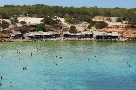 Formentera, Spain , Plaja de Cala Saona - July 21, 2015: Many people are swimming in a beautiful green sea, the fishing boats in the background under the characteristic huts in front of a pine forestのeditorial素材