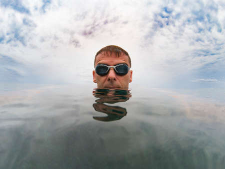 Young serious man in the sea, his head is on the smooth surface of the water, over him the cloudy sky, he wears gogglesの写真素材