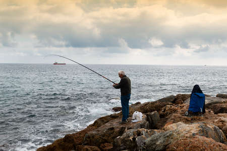 A fisherman standing with the fishing rod curved in his hand, he is on the rocks while trying to recover a fish, on the bottom an oil tanker sails on the horizon, the sky is cloudyの写真素材