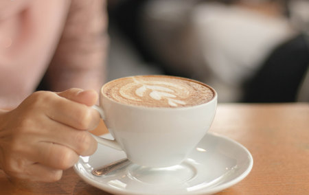 Closeup image of a hand holding a cup of hot latte coffee with blurry room on table at cafe coffee shop.の写真素材