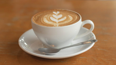 White cup of art latte on a cappuccino coffee on wood background standing on a table in a cafe. Foam flower on top of the cup.Little spoon and plate.Close up coffee cup on wooden table.の写真素材