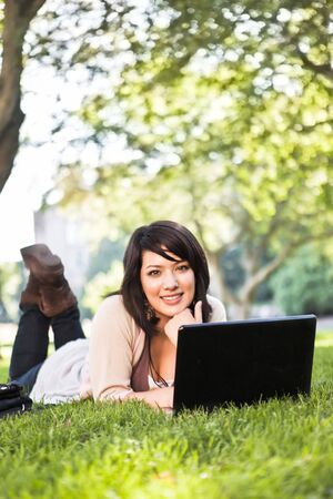 Mixed race college student lying down on the grass working on laptop at campusの写真素材