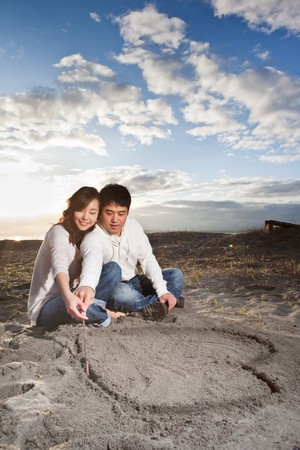 A portrait of an asian couple drawing love symbol on the sandの写真素材