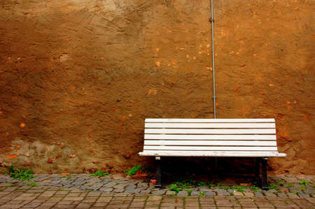 A single bench with white wooden stripes against a brown wall of an ancient houseの写真素材